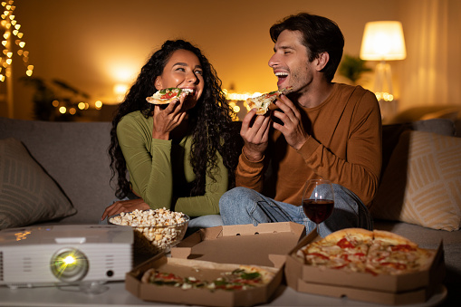 Joyful Couple Eating Pizza And Laughing Watching Comedy Film Using Home Cinema Projector Sitting On Couch Indoor. Entertainment And Fun Concept. Family Enjoying Movie On Weekend