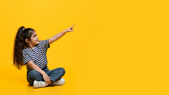 Check This. Adorable Little Arab Girl Sitting On Floor And Pointing Aside At Copy Space On Yellow Studio Background, Cheerful Cute Middle Eastern Female Child Recommending Something, Panorama