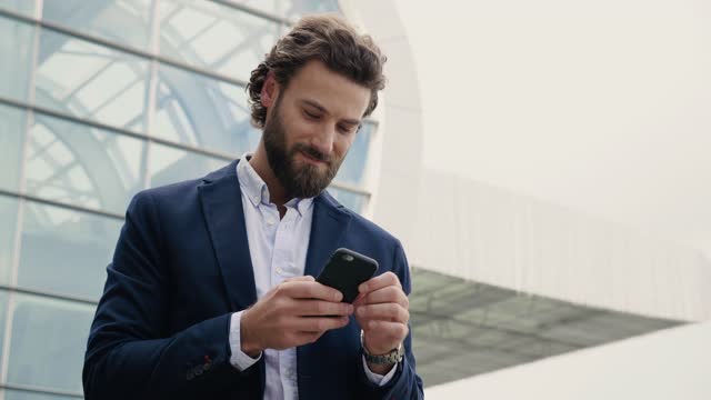 Handsome Businessman browsing his Smartphone near modern Office Building. Attractive Man is smiling and wearing Smart-casual Style. Social Networking. Luxury Lifestyle. Smartphones. Apps