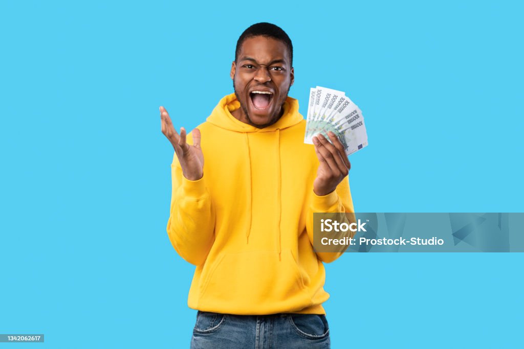 Emotional Black Man Shouting Holding Euro Money Over Blue Background Omg, I'm Rich. Emotional Black Man Shouting Holding Euro Money Cash Looking At Camera Standing Over Blue Studio Background. Profit And Wealth, Big Financial Luck And Success Concept Currency Stock Photo