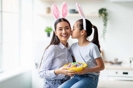 Holiday and celebration concept. Cute asian girl and woman wearing rabbit bunny ears on Easter day, copy space. Pretty kid kissing mom in cheek and holding basket full of painted eggs