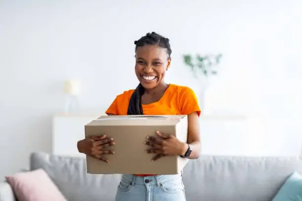 Photo of Overjoyed black lady holding cardboard parcel, receiving desired delivery, getting her online order at home
