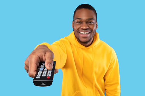 Cheerful Black Guy Watching TV Pointing Television Remote Controller To Camera Switching Channel Over Blue Studio Background. Happy Man Enjoying Movie. Selective Focus