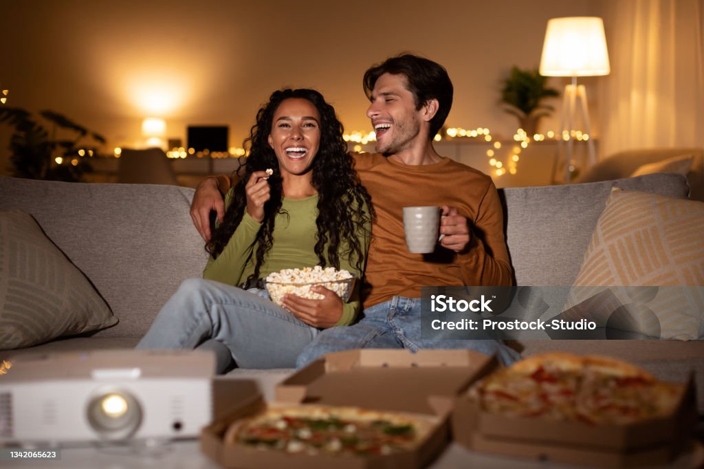 Cheerful Couple Using Home Cinema Projector Watching Comedy Movie Indoors Cheerful Couple Using Home Cinema Projector Watching Comedy Movie Laughing, Eating Popcorn And Pizza Sitting On Sofa Indoors. Weekend Family Leisure, Entertainment And Fun Movie Stock Photo