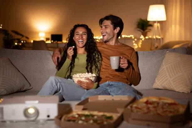 casal alegre usando projetor de cinema caseiro assistindo filme de comédia dentro de casa - family sofa night indoors - fotografias e filmes do acervo