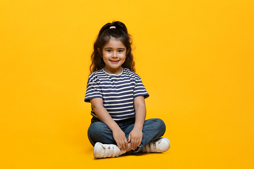 Portrait Of Adorable Little Middle Eastern Girl Posing Over Yellow Background, Cute Small Arab Female Child In Casual Clothes Sitting With Legs Crossed And Smiling At Camera, Copy Space