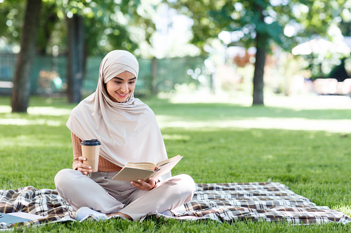 Smart hobby, leisure in free time, coffee break outdoor. Smiling young beautiful arab woman student in hijab sits on blanket on grass with cup of hot drink takeaway and reads book in park in summer