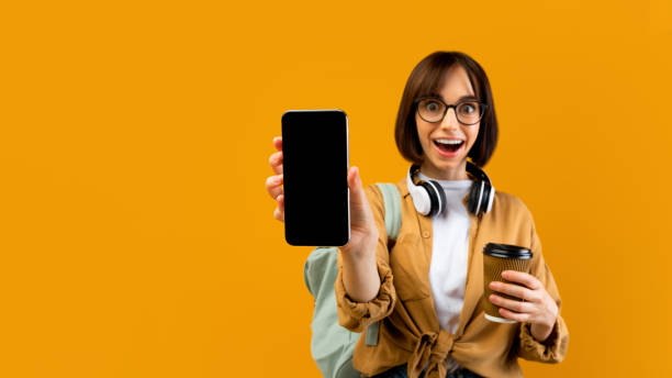 istock Emotional female student showing smartphone blank screen, posing with backpack and coffee on yellow background 1342067141