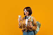 Happy female student with backpack, headphones, smartphone and takeaway coffee standing over yellow background