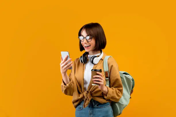 Happy female student with backpack, headphones, smartphone and takeaway coffee standing over yellow studio background. Young lady ready for classes, chatting on cellphone
