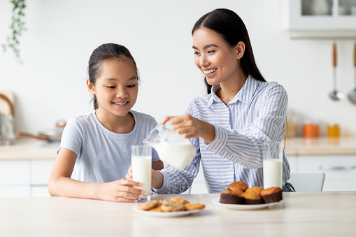 Happy asian mom and daughter eating snacks and drinking milk in kitchen interior. Young woman pouring calcium drink in glasses and having a bite together with little child girl