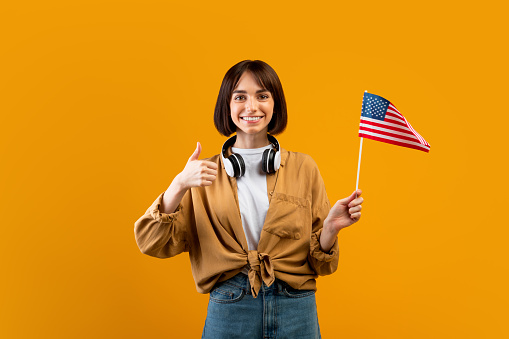 Happy lady showing thumb up and american flag, posing with headphones on neck over yellow studio background, copy space. Smiling female student with USA flag, education in America concept