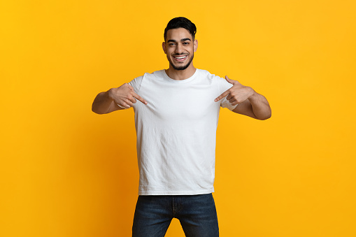 Cheerful arab guy pointing with both hands at his blank white t-shirt and smiling, posing over yellow studio background. Handsome middle-eastern young man showing something on his clothes, empty space
