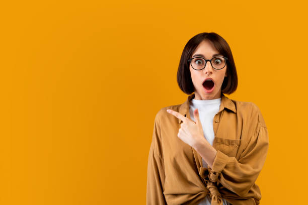 wow, look here. shocked lady pointing at empty space, showing place for advert or promotional text, yellow background - disbelief imagens e fotografias de stock