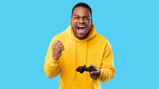 Gaming Concept. Emotional Black Guy Winning Video Game Posing With Joystick And Gesturing Yes In Joy Over Blue Studio Background. Millennial Man Celebrating Victory Playing Videogame. Panorama