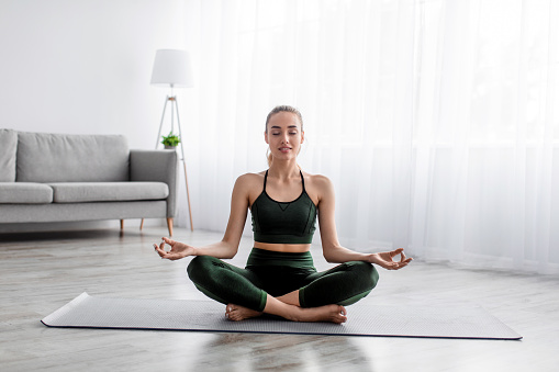 Calm serene european pretty millennial blonde lady practicing yoga, sitting in lotus position alone, female enjoys silence and peace at home interior, sun flare. Morning sports, body and health care