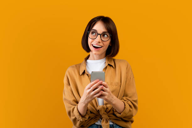 istock Dreamy young woman using smartphone and looking aside, standing over yellow studio background 1342073677