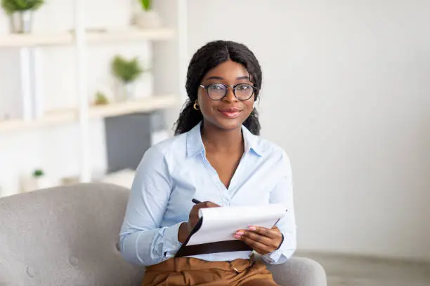 Photo of Friendly black female psychologist writing in clipboard, having session with client, sitting on couch at modern office