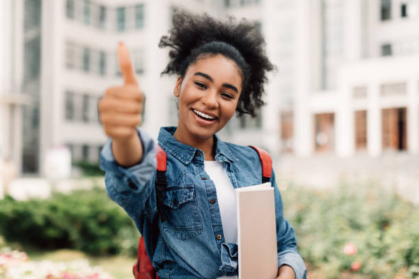 feliz estudante negra gesticulando polegares-up segurando livros posando do lado de fora - african ethnicity standing college student curly hair - fotografias e filmes do acervo