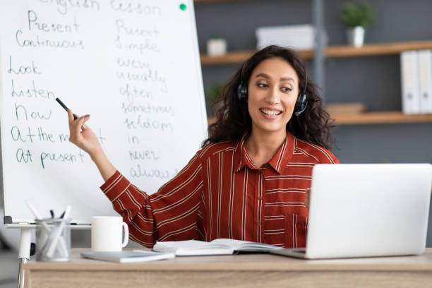 Confident English teacher showing grammar rules to students online Elearning Concept. Portrait of smiling female teacher having video conference call using laptop. Lady in wireless headphones talking to webcam, recording tutorial on pc for students pointing at board online tutors stock pictures, royalty-free photos & images