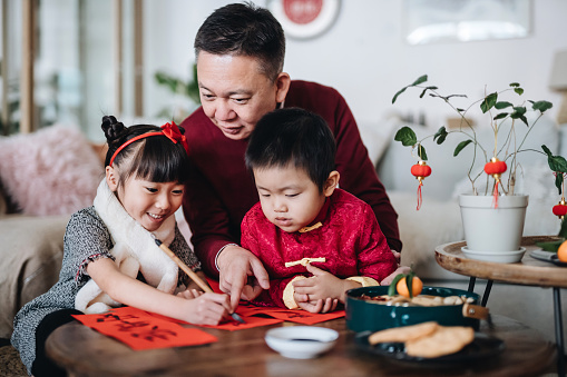 Joyful little granddaughter and grandson practising Chinese calligraphy for Chinese New Year Fai Chun (Auspicious Messages) on couplets with their grandfather at home