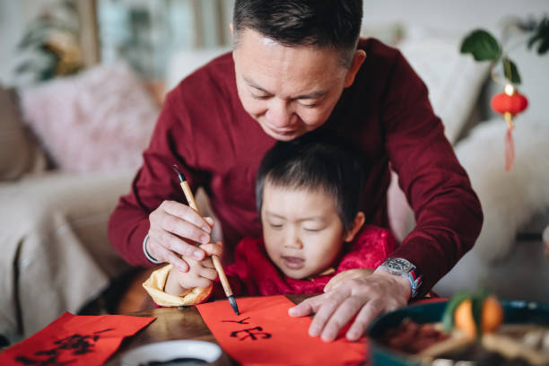 Grandfather practising Chinese calligraphy for Chinese New Year Fai Chun (Auspicious Messages) and teaching his grandson by writing on couplets at home stock photo