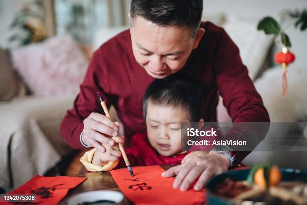 Grandfather Practising Chinese Calligraphy For Chinese New Year Fai Chun And Teaching His Grandson By Writing On Couplets At Home Stock Photo - Download Image Now