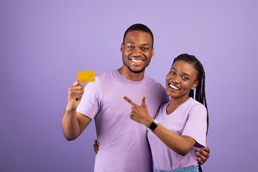 Happy Black Couple Holding And Pointing Finger At Credit Card Recommending Service Smiling To Camera Over Purple Background. Excited Bank Customers Showing Card For Payments Posing In Studio