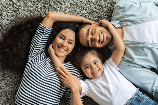 Closeup portrait of happy middle-eastern family posing on floor at home, laying on carpet and bonding, cheerfully smiling at camera, view above. Positive father, mother and daughter cuddling, top view