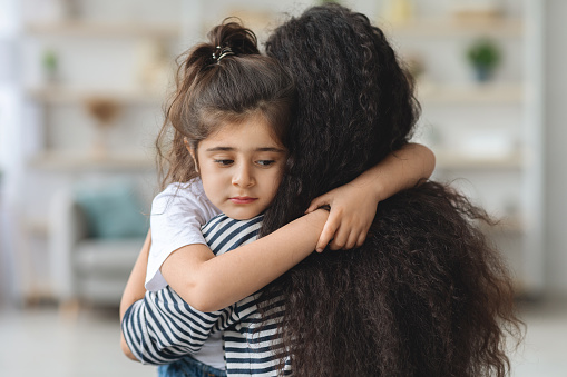 Upset little girl hugging her mom, home interior, closeup portrait, copy space. Long-haired middle-eastern mother comforting her sad crying daughter. Motherhood, mother love concept