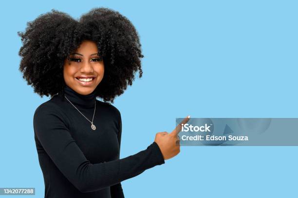 Portrait Of A Smiling Young Afro Girl Points To The Rigth Side Isolated On A Blue Background Stock Photo - Download Image Now