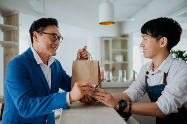 Asian man using credit card contactless payment at cafe Image of an Asian Chinese man paying with credit card contactless payment to cafe store worker. Credit card contactless wave payment, cashless transaction at cafe. asian cashier stock pictures, royalty-free photos & images