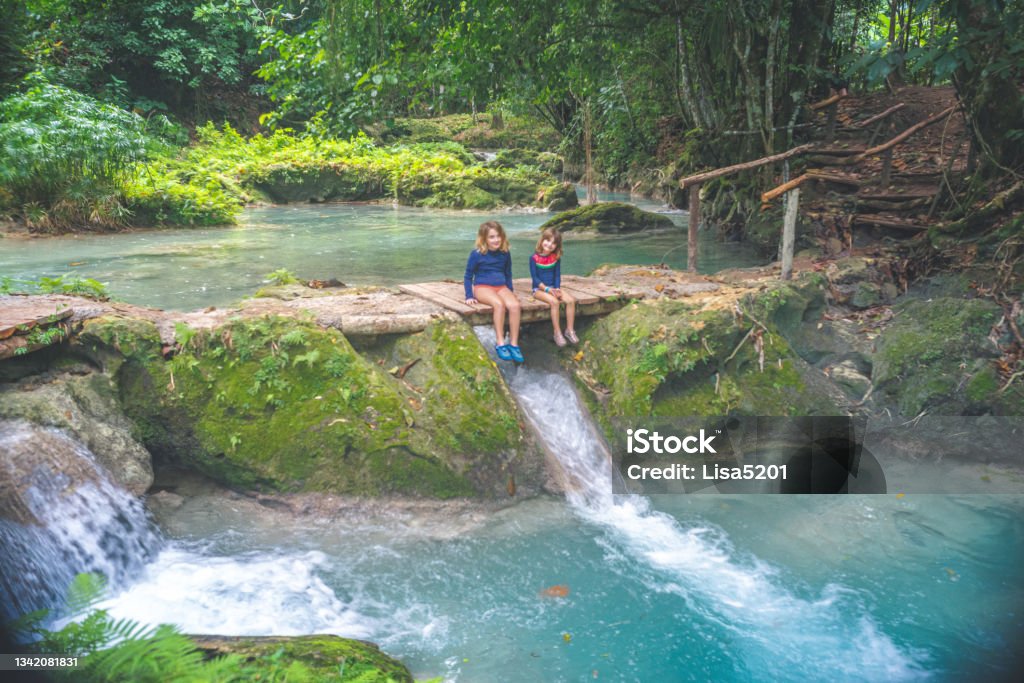 Children sit by a lush tropical lagoon waterfall on a family vacation Adventurous kids by a waterfall Jamaica Stock Photo