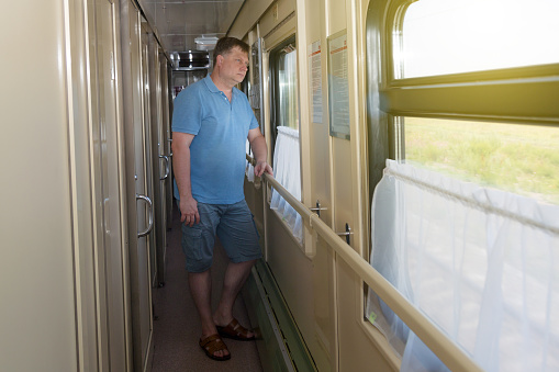 A blond man of 35-40 years old is standing in corridor of the train and looks out the window.