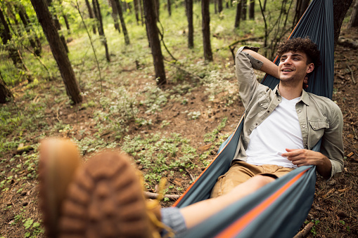 Cheerful young Caucasian male hiker resting in a hammock in the pine forest.