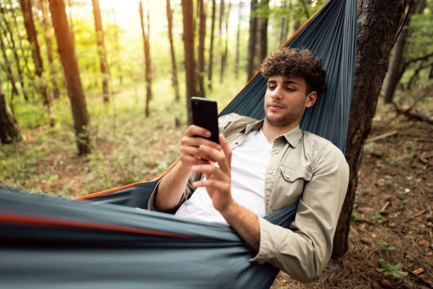 Cheerful male hiker relaxing in a hammock in the pine forest and using a smart phone stock photo