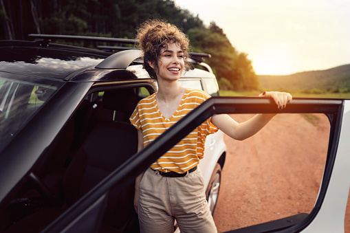 Beautiful young Caucasian woman with curly hair standing by the car and looking away. Woman going on a summer road trip.