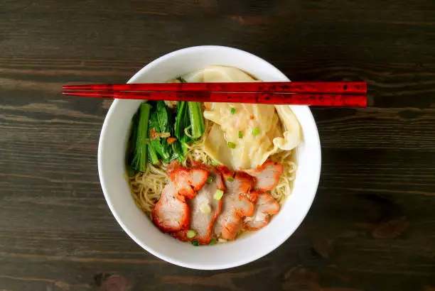 Photo of Bowl of Egg Noodle and Wonton Dumpling Soup with Roasted Pork Isolated on Wooden Table