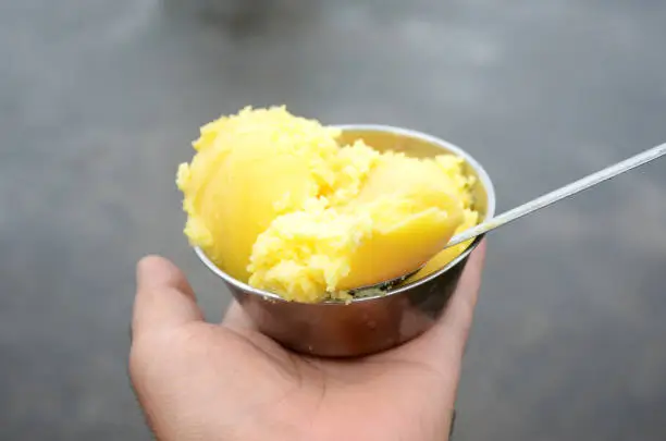 Photo of closeup the yellow clarified butter in the steel bowl with steel spoon hold hand over out of focus brown background.