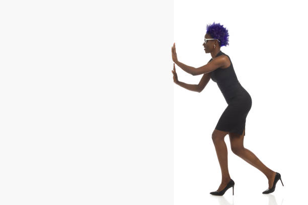 mulher afro-americana elegante de salto alto e mulher americana arficana negra em mini vestido preto está empurrando bandeira branca vazia. vista lateral. tiro de comprimento completo. - pushing women wall people - fotografias e filmes do acervo