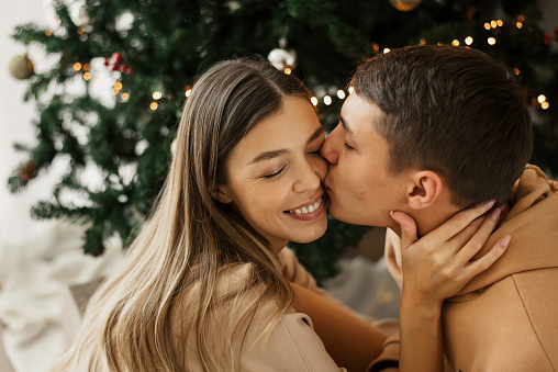 Young couple sitting near Christmas tree at home. Man kissing a woman. Love and relationships. Christmas and New Year celebration