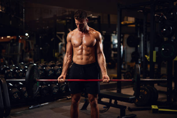 Muscular shirtless male bodybuilder lifting a barbell in a gym stock photo