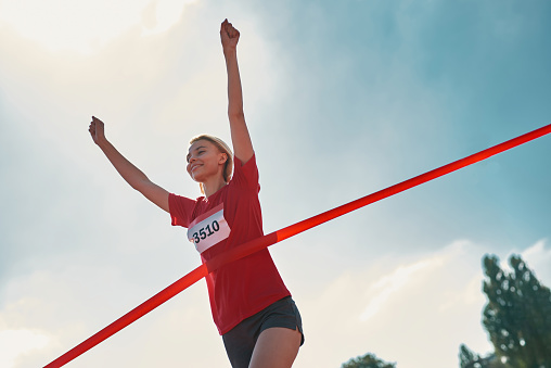 Low angle view of happy young female runner with arms raised reaching the finish line at track field during marathon outdoors. Sports, motivation concept