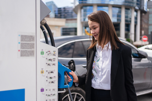 Young woman charging an electric car at public charging station and pays using a mobile phone. Innovative eco-friendly vehicle