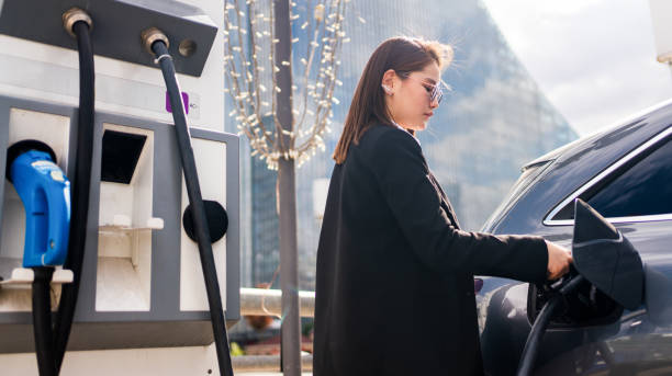 giovane donna che carica un'auto elettrica alla stazione di ricarica pubblica e paga usando un telefono cellulare. veicolo ecologico innovativo - gas station foto e immagini stock