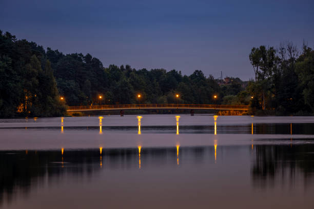 The bridge on the Dlugie Lake is illuminated by the light from the lamps after sunset. The light from the lamps is reflected in the water and leaves long yellow streaks. - Olsztyn, Warmia and Mazury, Poland