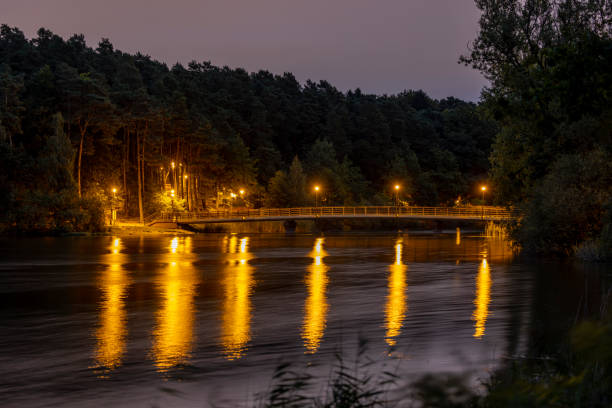 The bridge on the Dlugie Lake is illuminated by the light from the lamps after sunset. The light from the lamps is reflected in the water and leaves long yellow streaks. - Olsztyn, Warmia and Mazury, Poland