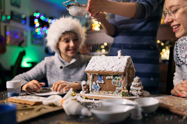 Mother and kids enjoying building and decorating gingerbread house for Christmas. The mother is dusting powdered sugar on the house. It looks like it's snowing.\nCanon R5.