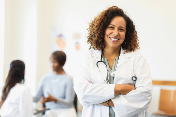 Portrait of confident mid adult female doctor A confident female doctor smiles at the camera while standing with her arms crossed. A colleague and a patient are talking in the background. general practitioner stock pictures, royalty-free photos & images