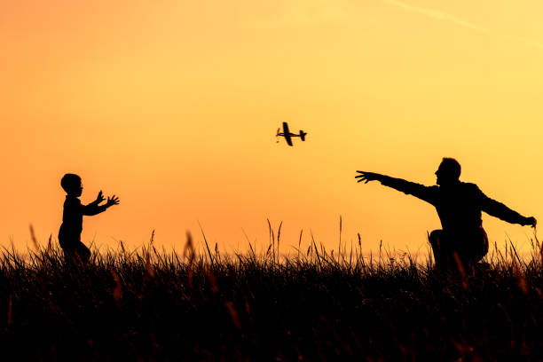 силуэты отца и маленького сына, играющих с игровым самолетиком на фоне заката. - catch light стоковые фото и изображения